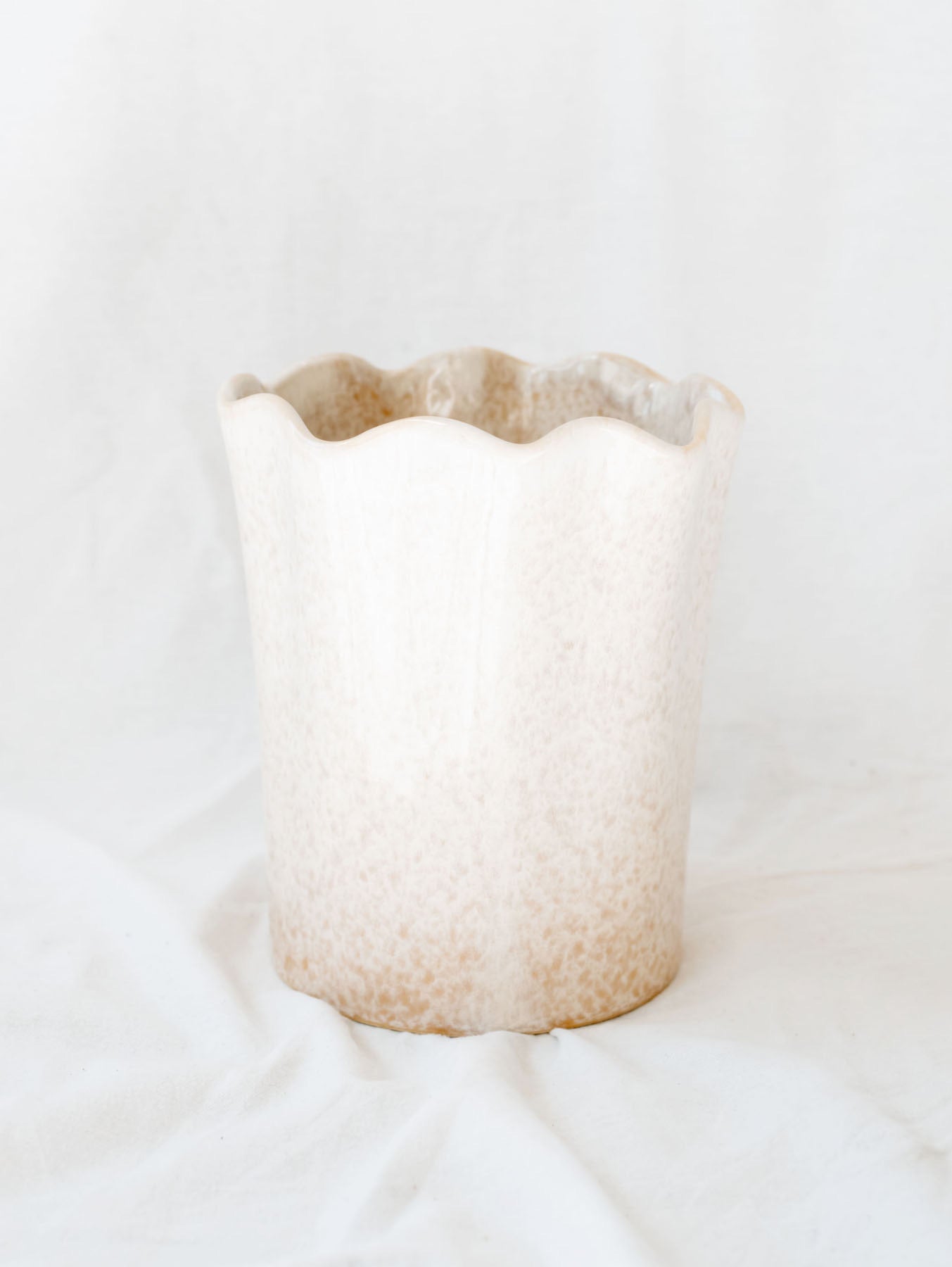 Beige scallop edge large mouth short vase standing against white linen backdrop. 