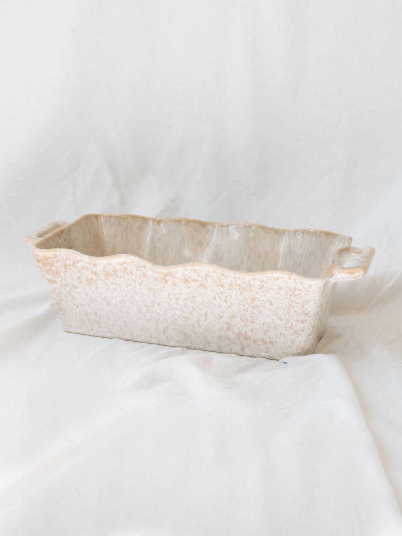 Side view of beige scallop edge baking dish on white linen backdrop.