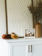 Velvet pumpkins styled on a shelf with large textured artwork, a small seasonal art pieces, and a warm wood vase with a thistle faux foliage stem. 
