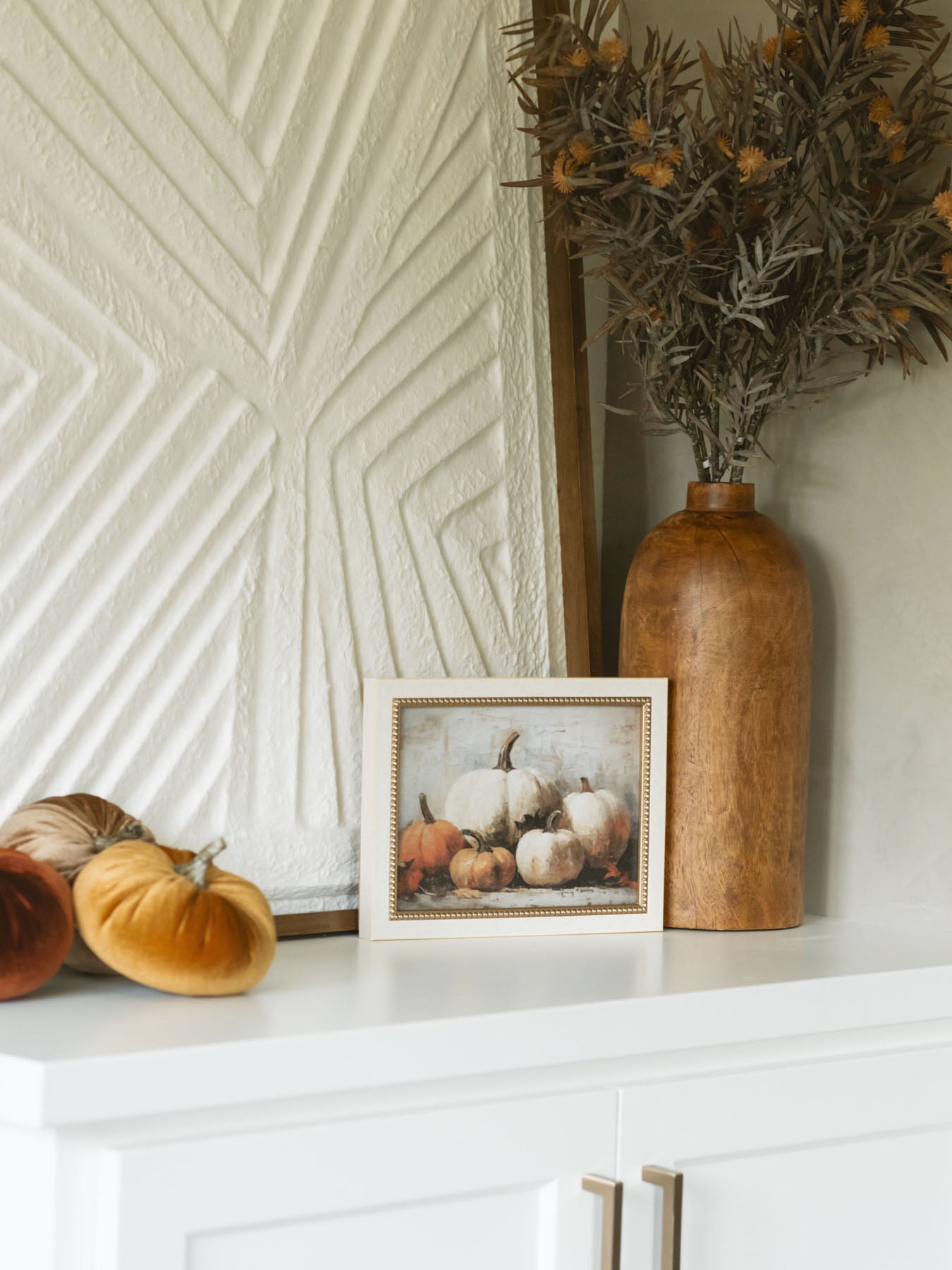 Velvet pumpkins styled on a shelf with large textured artwork, a small seasonal art pieces, and a warm wood vase with a thistle faux foliage stem. 