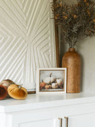 Velvet pumpkins styled on a shelf with large textured artwork, a small seasonal art pieces, and a warm wood vase with a thistle faux foliage stem. 