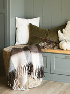 Brown check throw blanket with fringe edges layered into a basket in front of a bench with pillows and soft fabric pumpkins. 