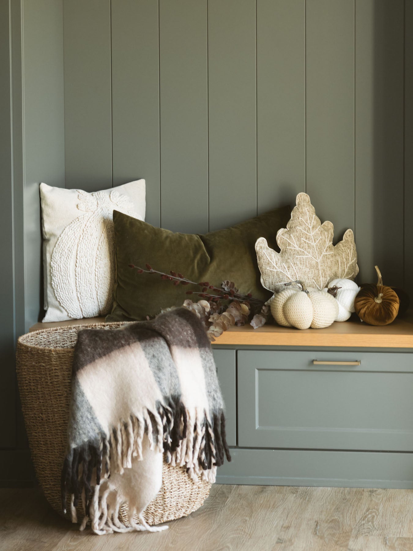 Brown check throw blanket with fringe edges layered into a basket in front of a bench with pillows and soft fabric pumpkins. 