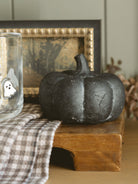 The smallest size in the pumpkin shape is styled sitting on a warm wood board with a dark framed fall inspired art piece and a clear drinking glass with embedded ghosts on it. 