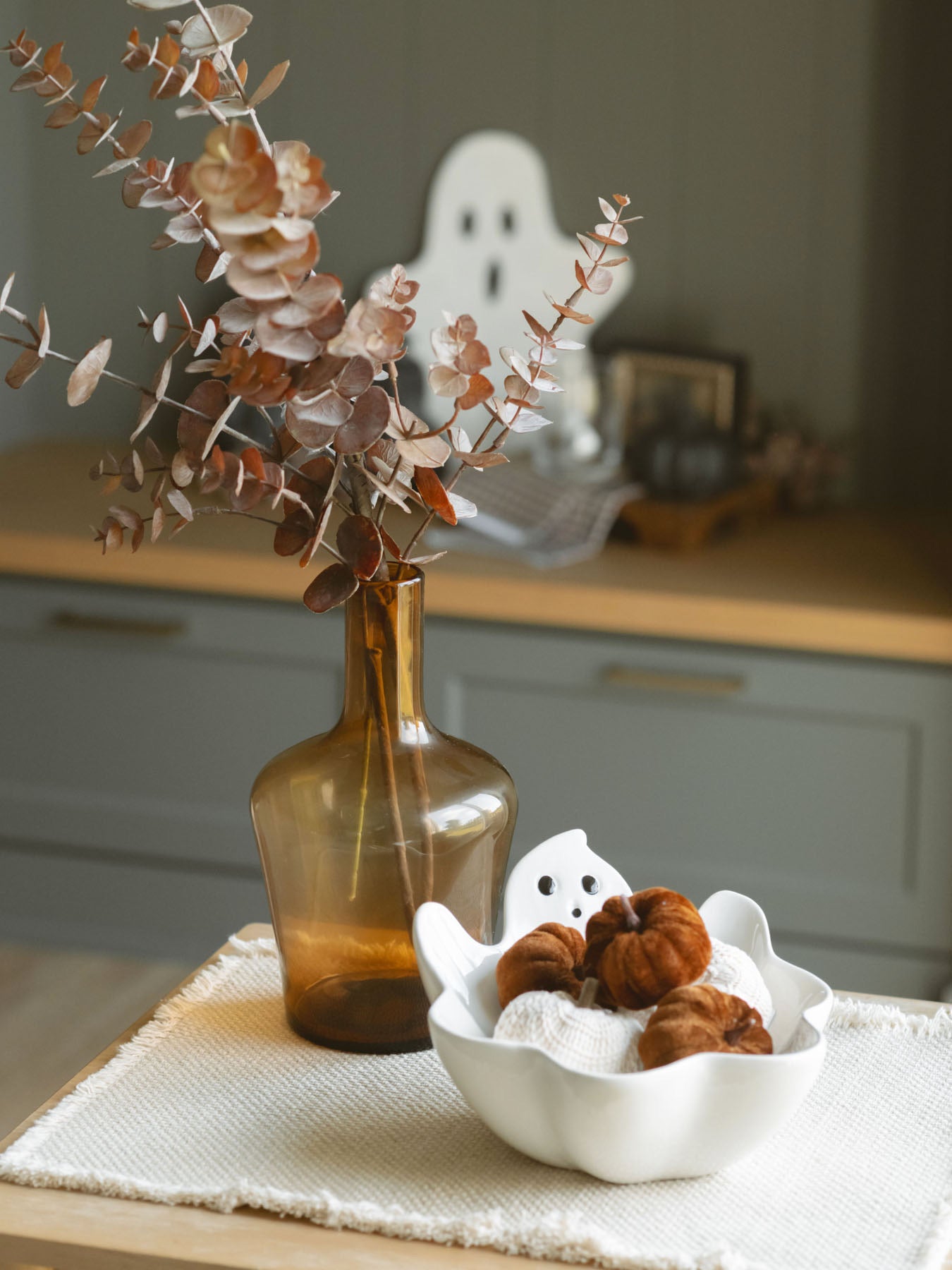 Amber glass vase filled with a brown eucalyptus layered with the white ghost bowl and a beige placemat. 