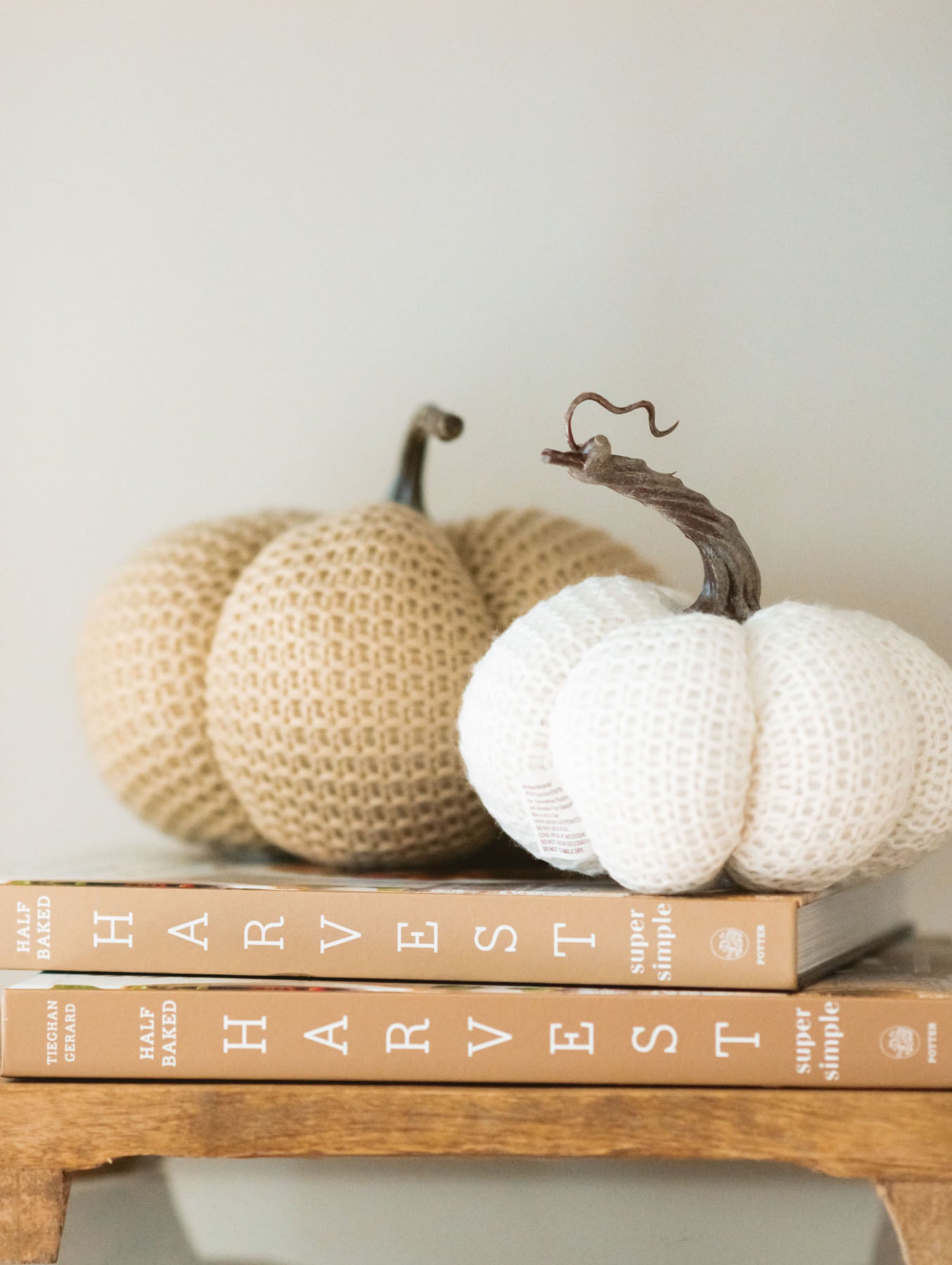 Tan knit stuffed pumpkin with medium sized white knit stuffed pumpkin styled on some books and a wood riser board.