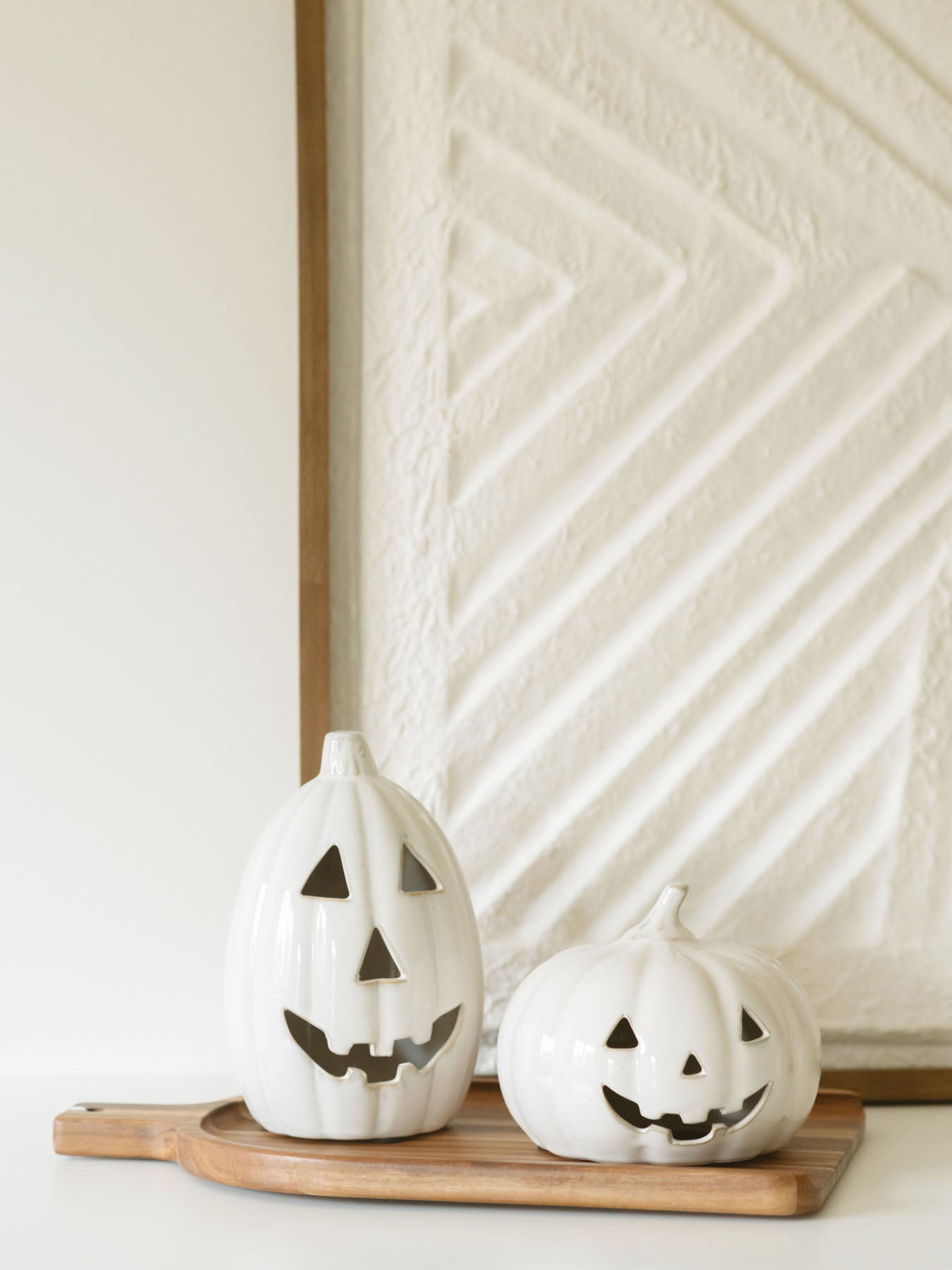 The two sizes of the white ceramic jack-o-lanterns sitting next to each other on a warm wood board, set in front of a large textured piece of artwork. 