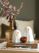 Two sizes of the white ceramic ghosts sitting next to each other on a warm wood riser with a small brown soft pumpkin and the amber glass vase with brown eucalyptus. 