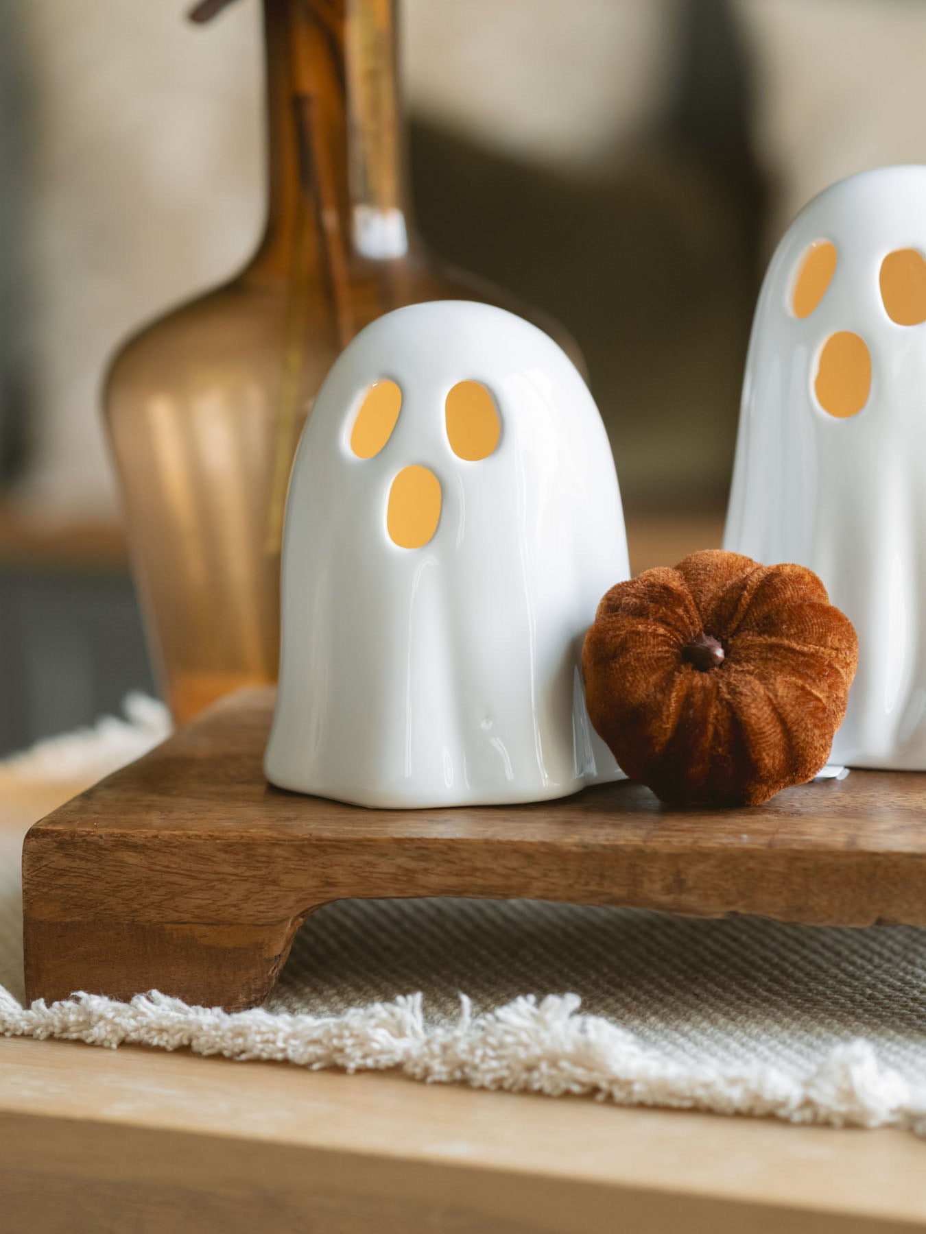 Two sizes of the white ceramic ghosts sitting next to each other on a warm wood riser with a small brown soft pumpkin and the amber glass vase with brown eucalyptus. 
