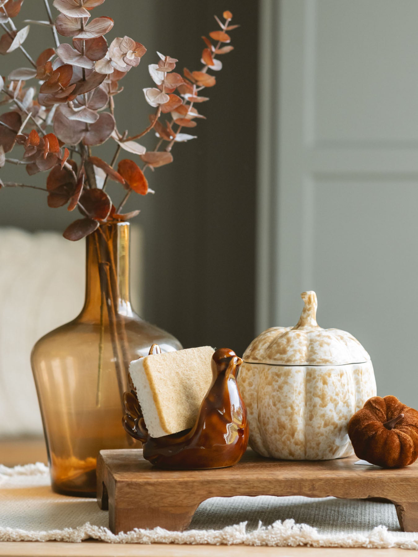 Stoneware pumpkin shaped jar with lid on top of it styled with warm brown tones. Layered onto a wood board with the stoneware turkey sponge holder, a mini velvet pumpkin, and an amber glass vase and sprigs to create a full fall moment. 