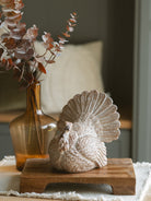Resin tabletop turkey sitting on a warm wood riser paired with the amber glass vase with a brown eucalyptus stem. 