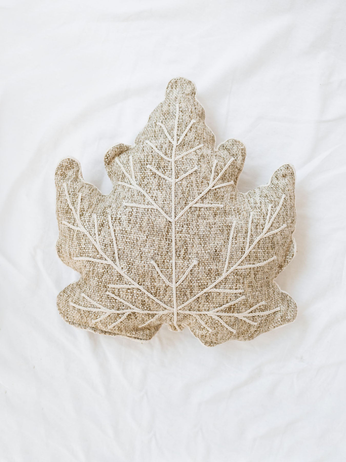 Oatmeal and sage tweed leaf shaped pillow laying on white linen backdrop. 