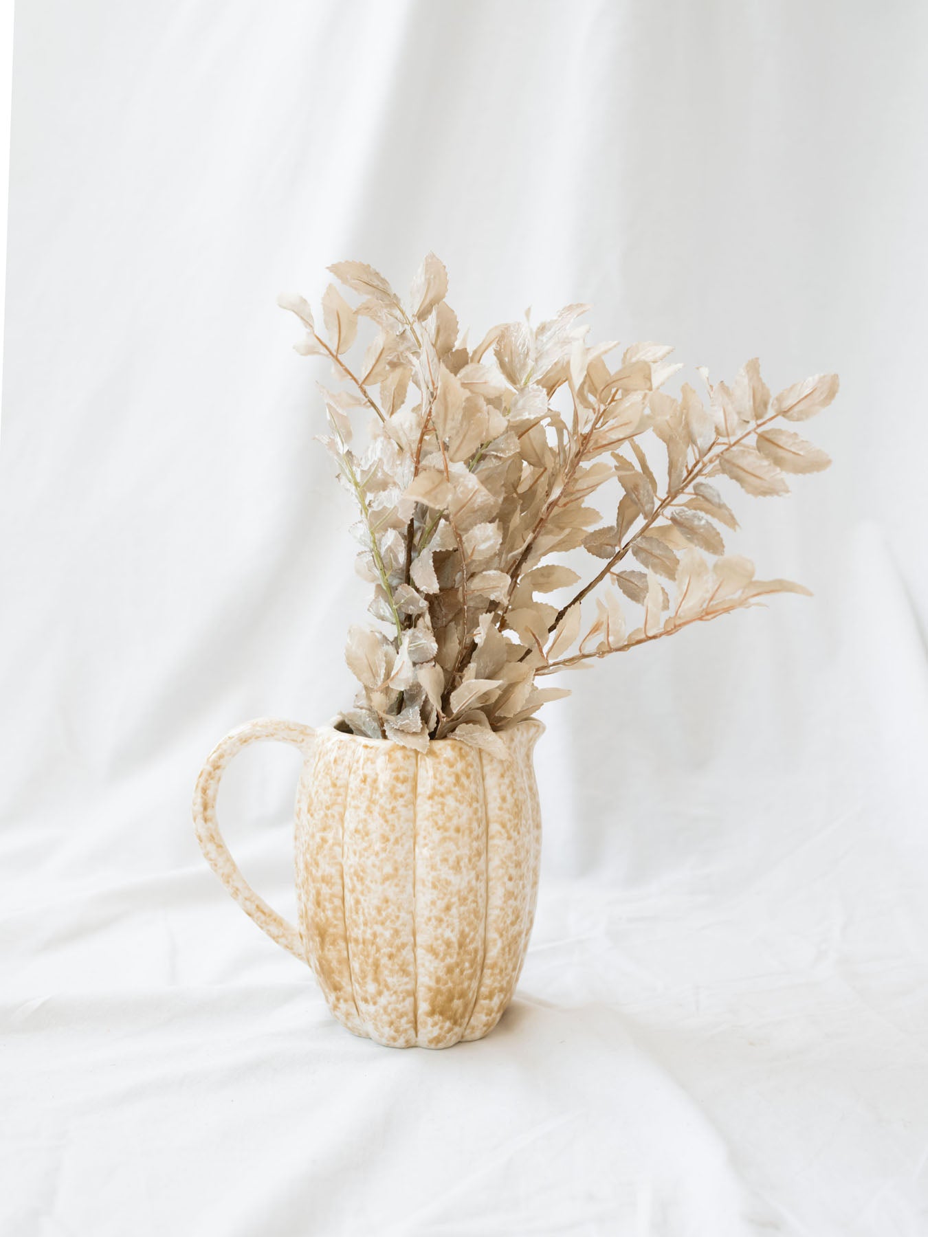 Showing the stoneware pitcher styled with a faux foliage in a cream color sitting on a white linen backdrop. 