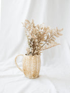 Showing the stoneware pitcher styled with a faux foliage in a cream color sitting on a white linen backdrop. 