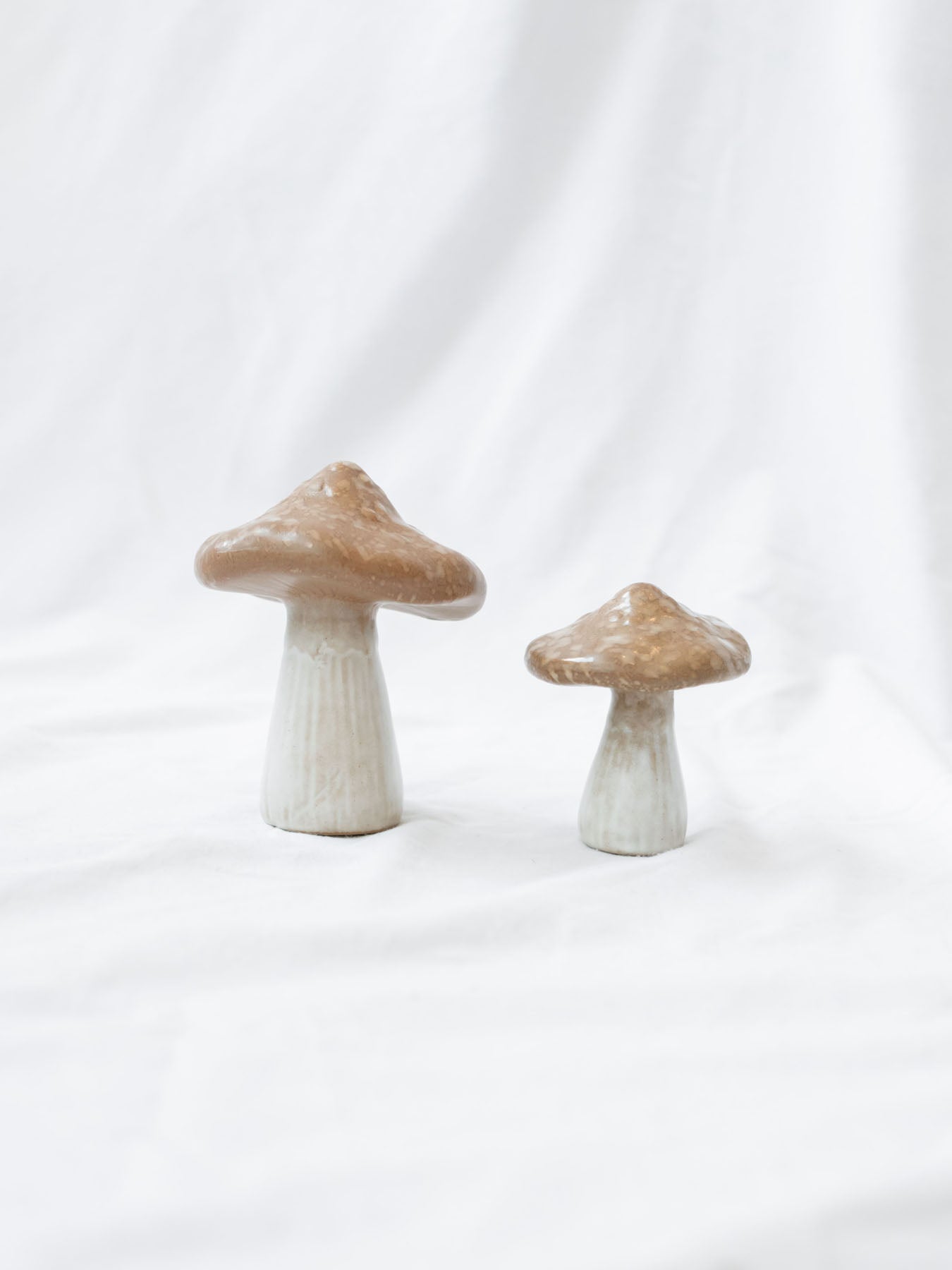 Both stoneware mushrooms sitting next to each other to see sizing difference, they are set up on a white linen backdrop. 