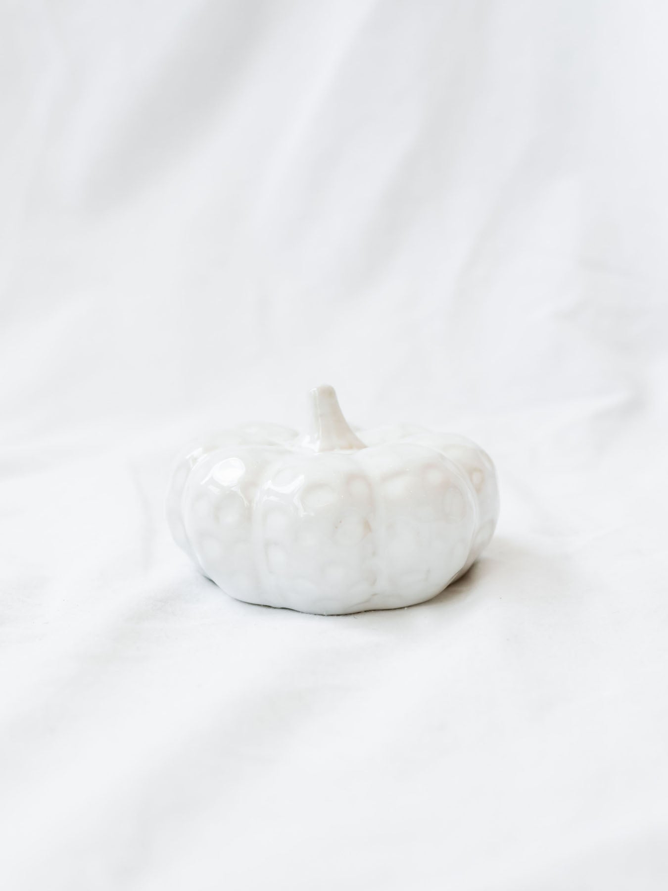 White ceramic textured pumpkin shown sitting on white linen backdrop. 