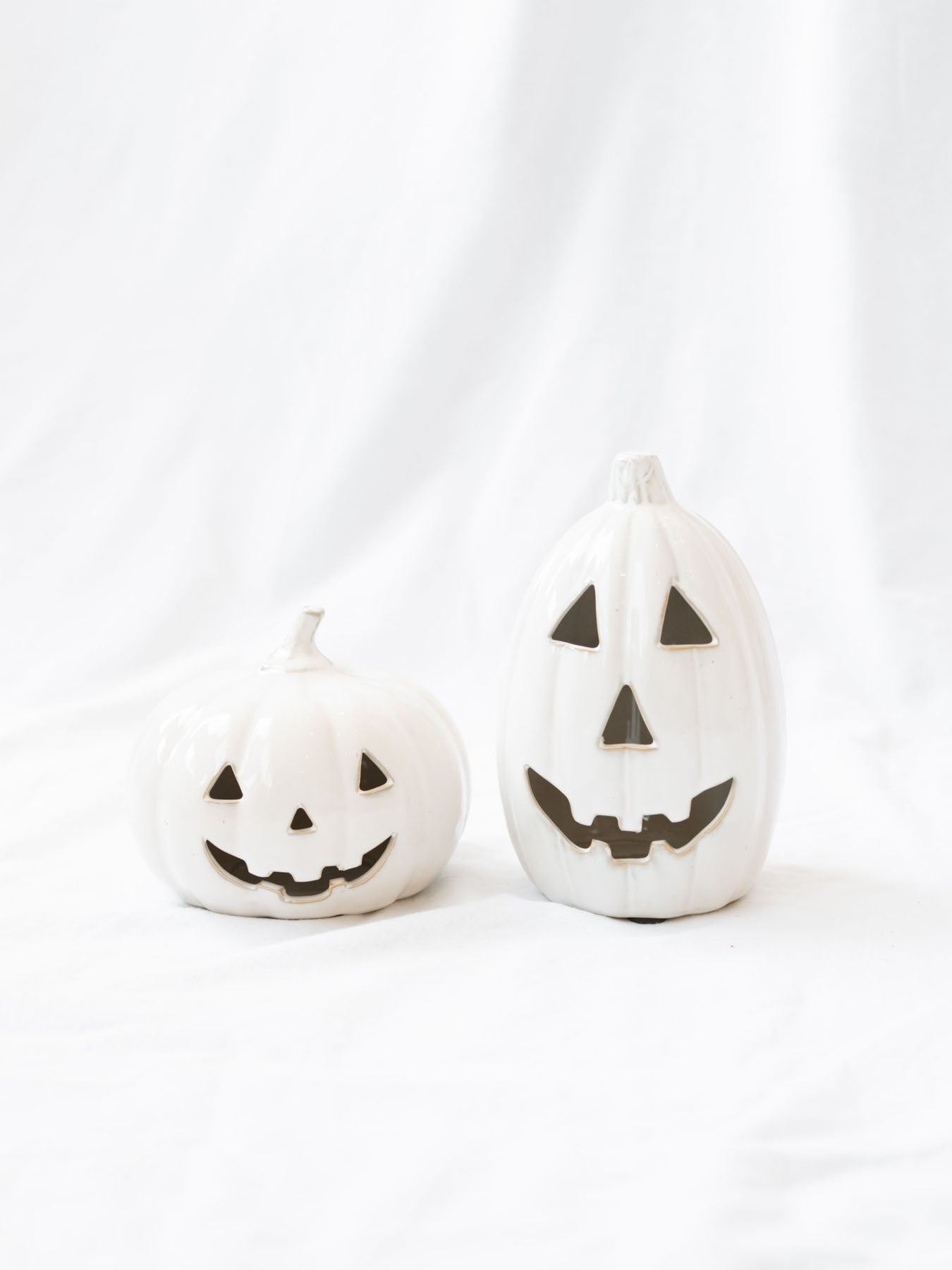 The two sizes of the white ceramic jack-o-lanterns sitting next to each other on a white linen backdrop. 