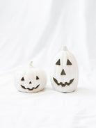 The two sizes of the white ceramic jack-o-lanterns sitting next to each other on a white linen backdrop. 