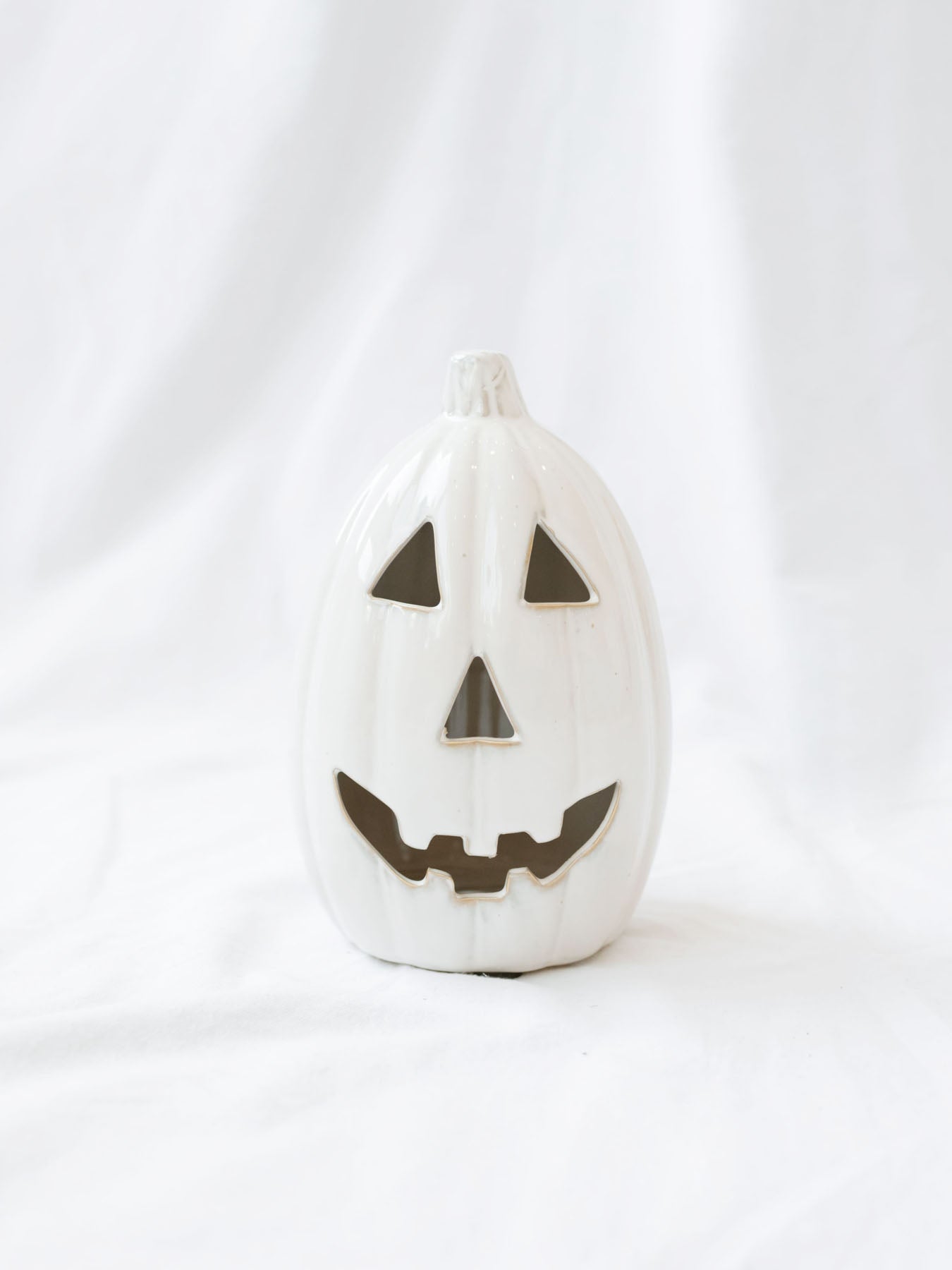 The large taller size of the white ceramic jack-o-lanterns sitting on a white linen backdrop. 