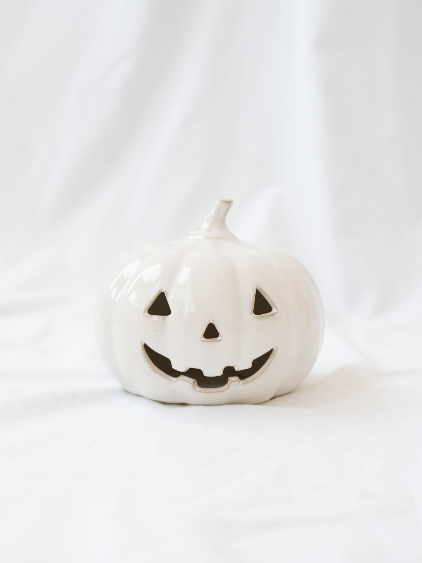The Small shorter size of the white ceramic jack-o-lanterns sitting on a white linen backdrop. 