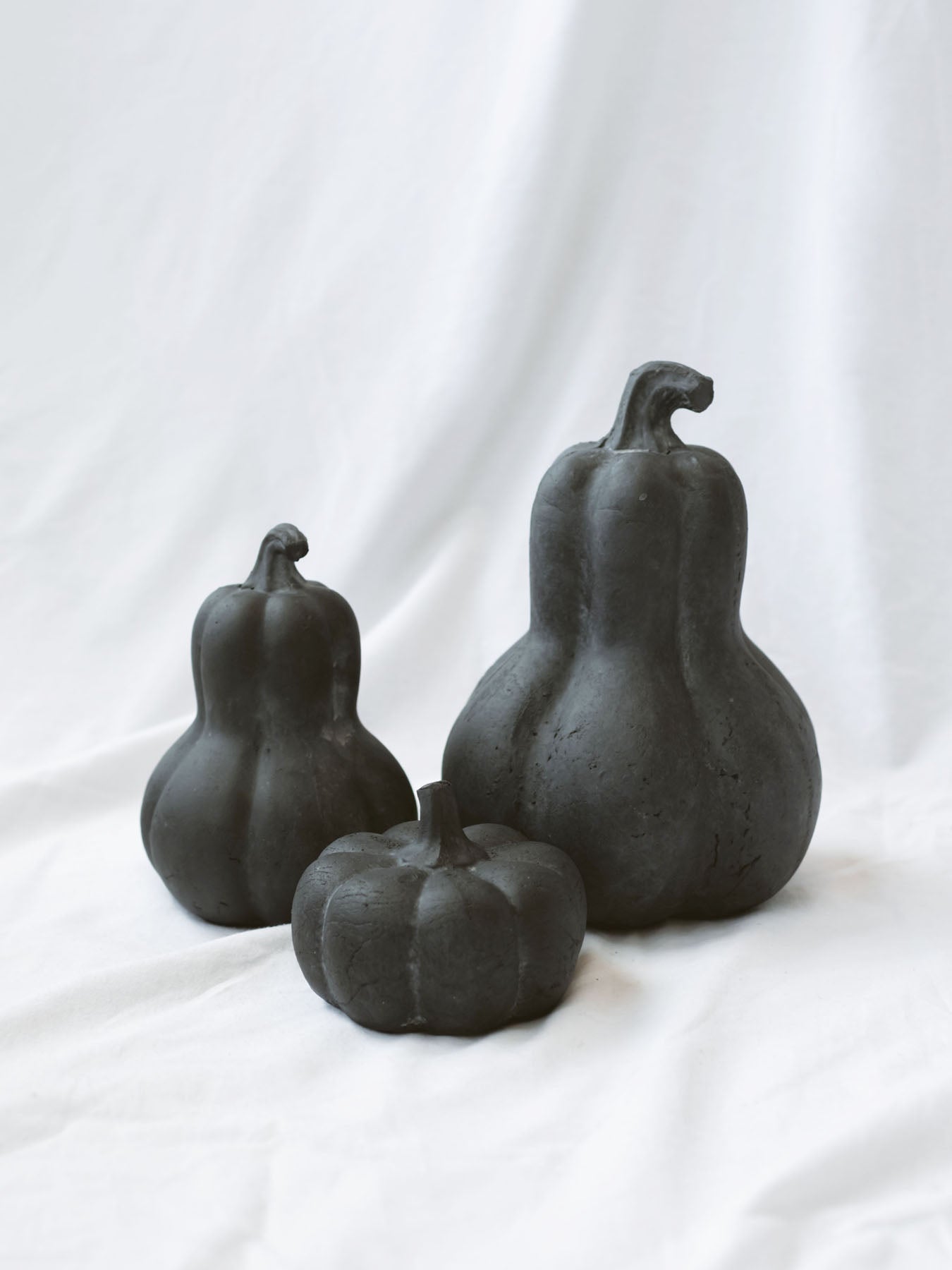 Two gourds and one pumpkin in a black terracotta. All three sizes are sitting on a white linen backdrop. 