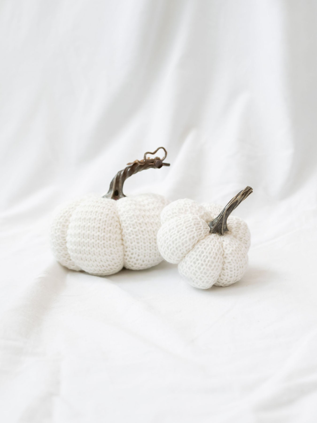 Small and large size knit stuffed white pumpkins on white linen backdrop. 