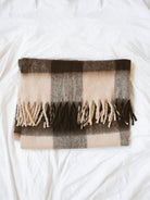 Brown check throw blanket with fringe edges folded laying on white linen backdrop. 