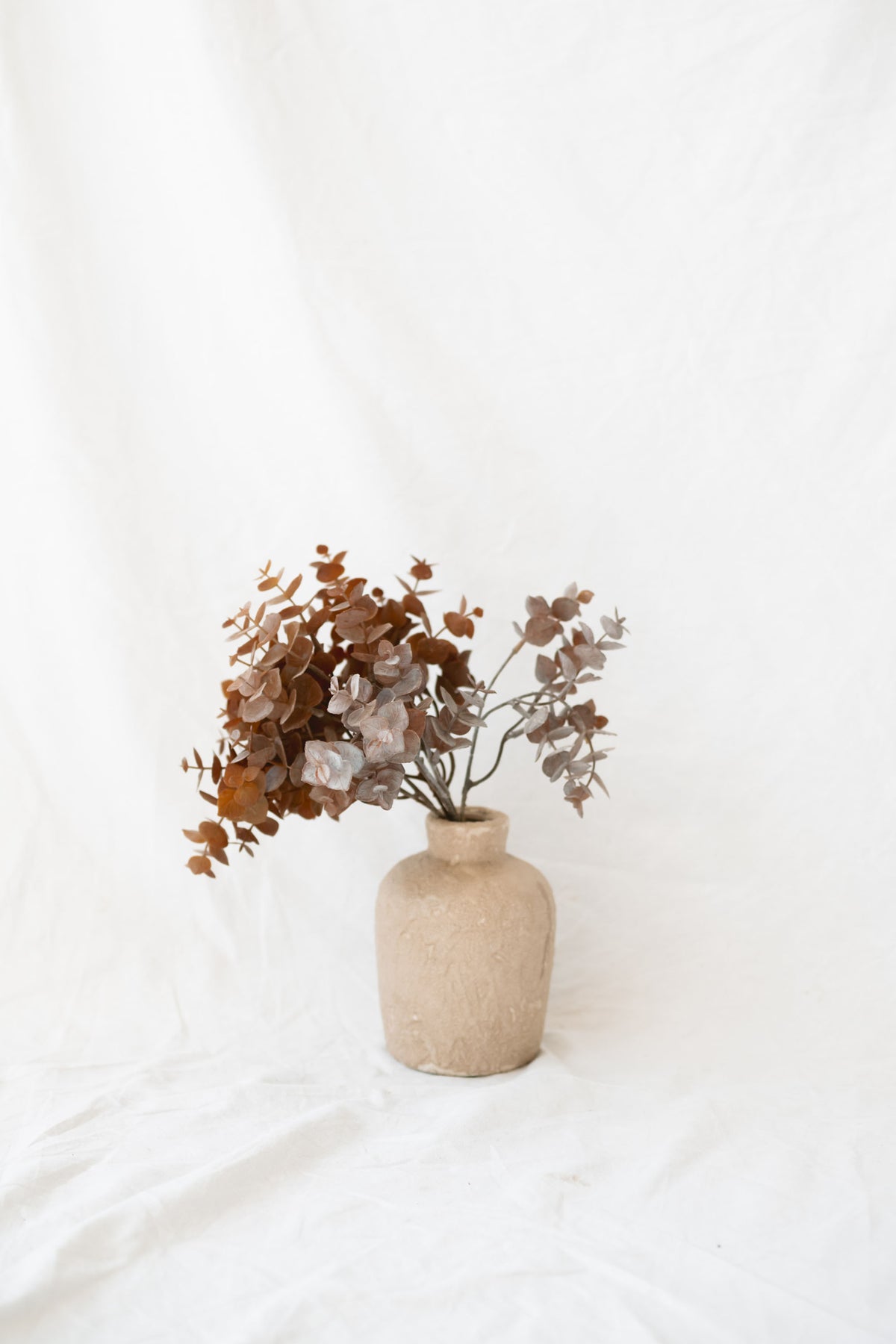 Brown eucalyptus bundle sitting in small vase against white linen backdrop.