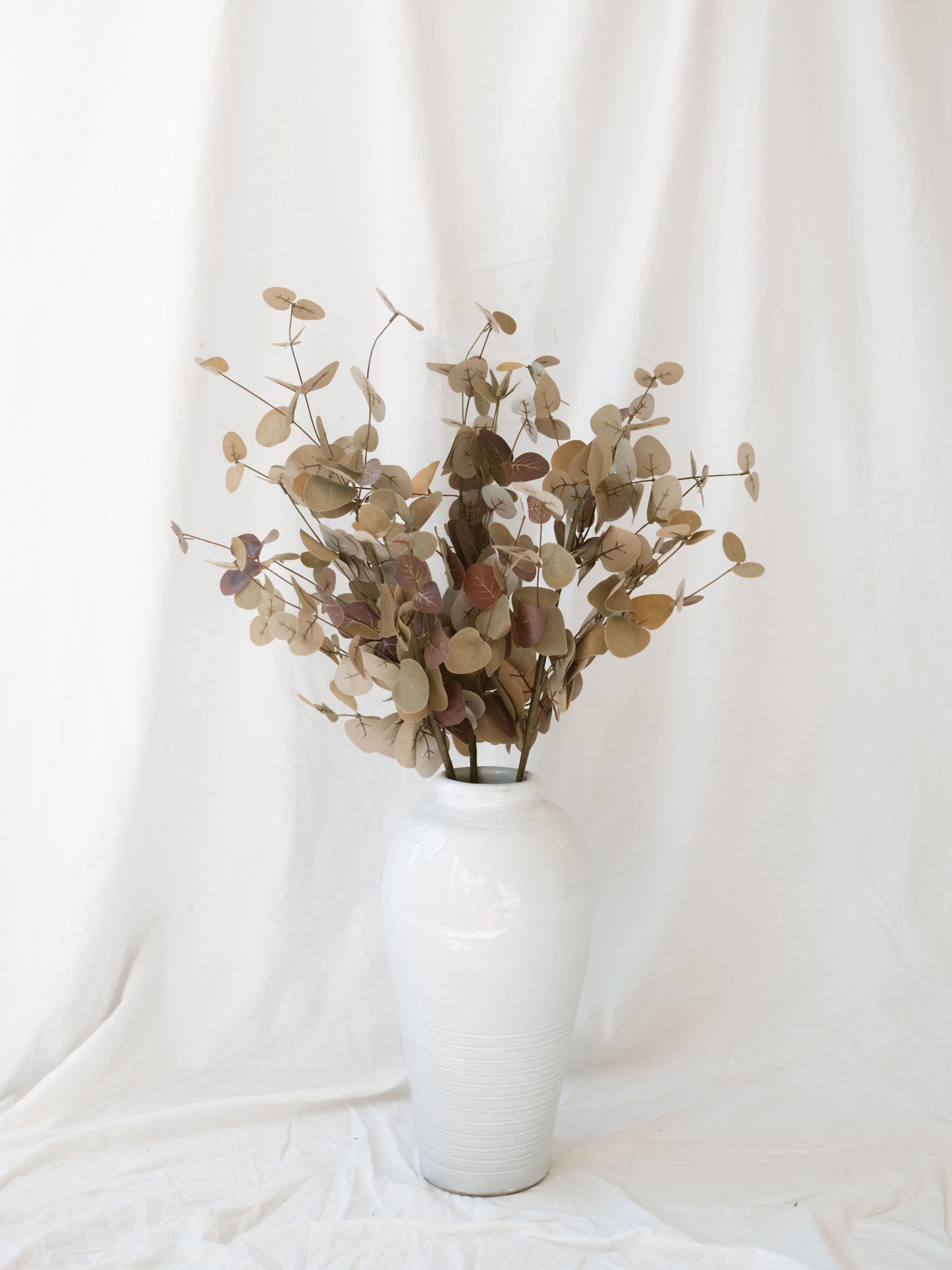 Two tone tan eucalyptus stem in white vase sitting in front of white linen backdrop.